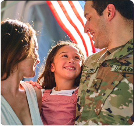 A happy military family of husband, wife, and child standing in the sunshine in front of the home decorated with an American flag